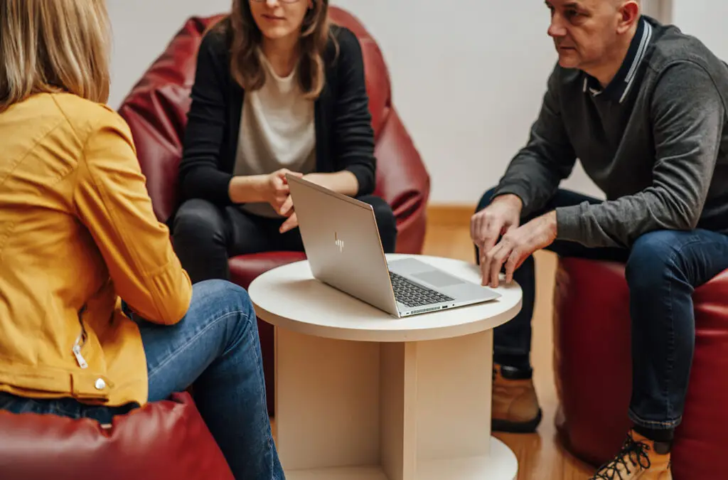 Trije sodelavci sedijo okoli ogrogle mizice, na kateri se nahaja računalni, in snujejo prenovo sistema nagrajevanja.