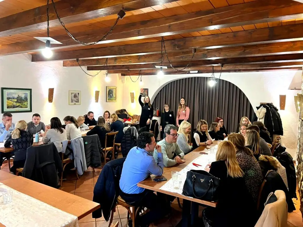 Skupina ljudi sedi za lesenimi mizami v prijetni sobi z lesenimi tramovi in belimi stenami, okrašenimi s slikami. Udeleženci delavnice razvoja talentov se pogovarjajo in sodelujejo.